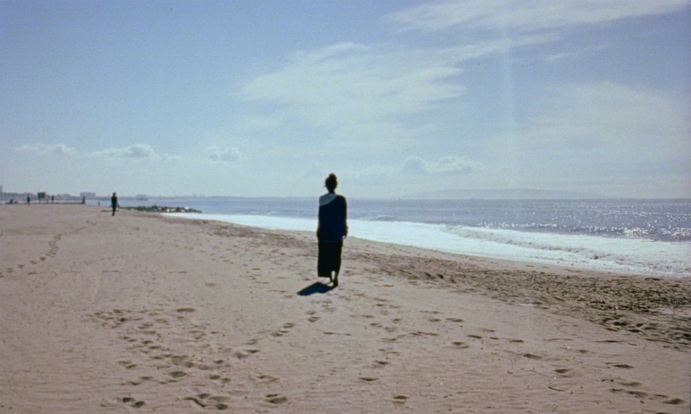 Landscape of a shoreline on a blue sunny day, footsteps visible in the sand and a figure whose back is turned toward the viewer.