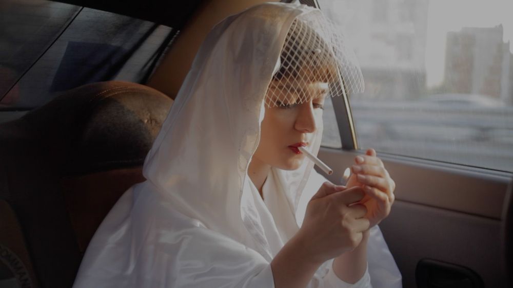 A person is seated in the backseat of a vehicle. Their attire consists of a white robe or jacket, paired with a delicate white birdcage veil that drapes over their hair and face. 