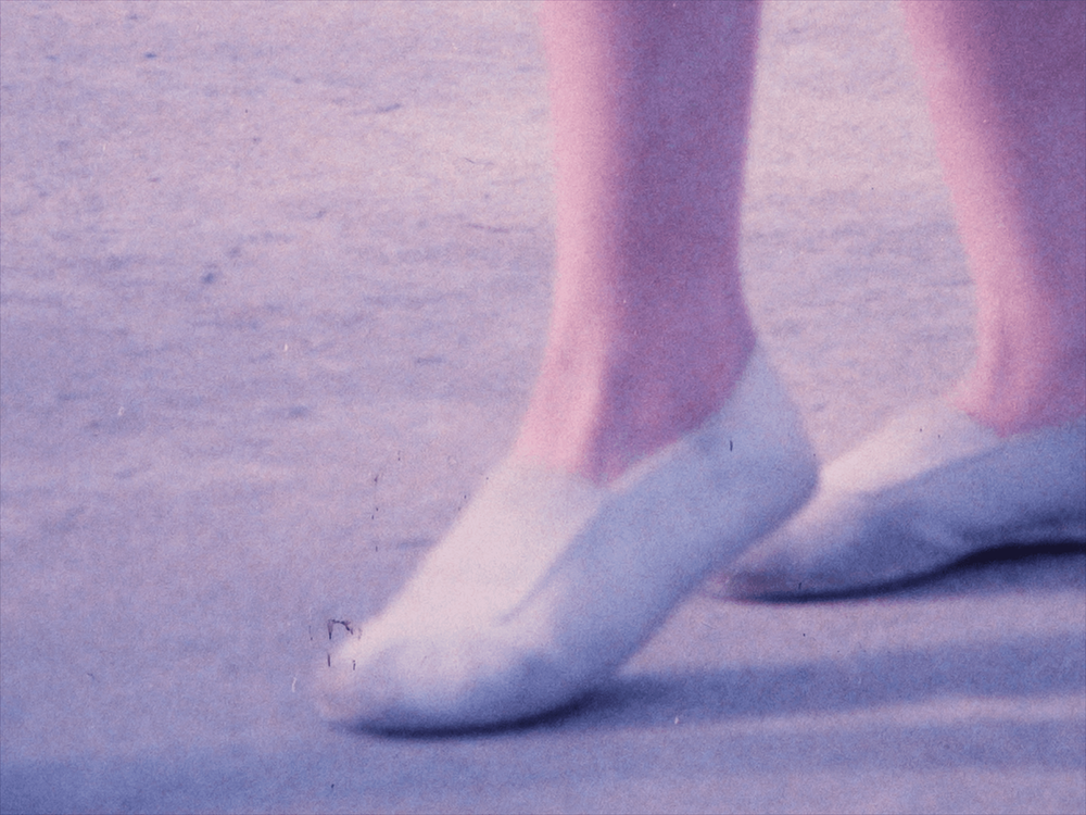 Blue-tinged close-up of a pair of feet wearing white flats, slightly tip-toeing on concrete.