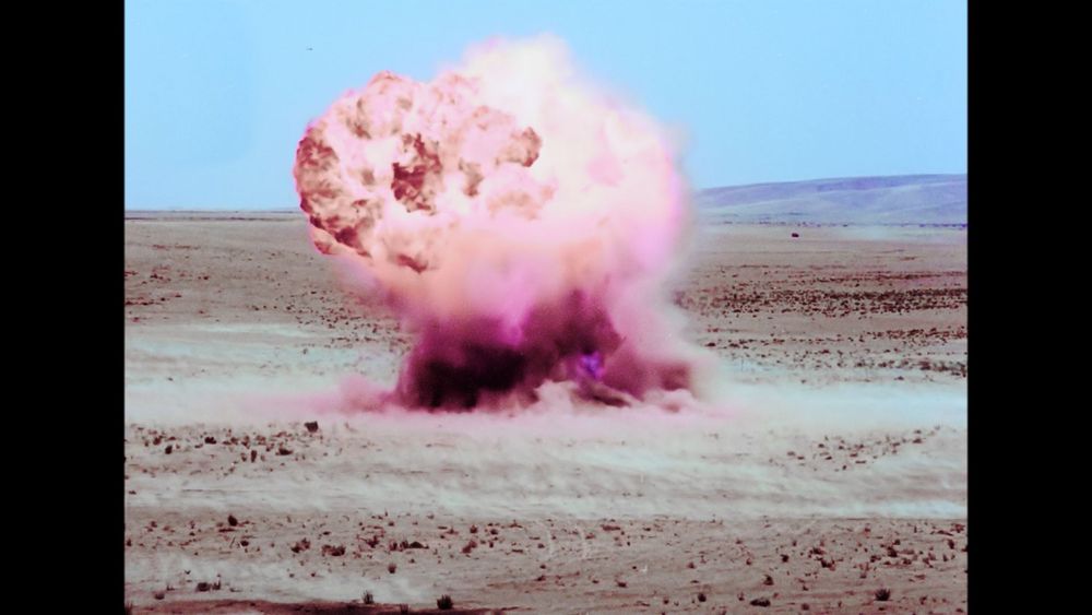 Sandy landscape on a blue, sunny day. There is an explosion coloured in pink and purple tones at the center of the landscape.
