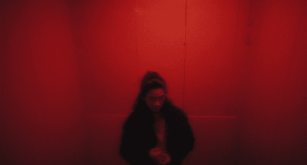 Hazy, small, red-walled room with a person standing in the middle, visible from the waist up. Their long black hair is in a half bun and they wear a black jacket with a white shirt underneath.