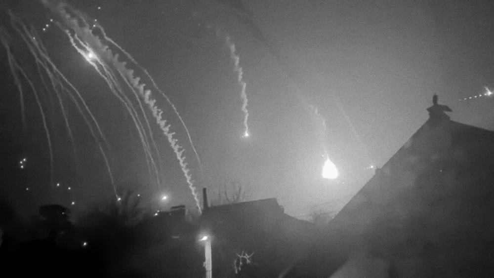 Black and white scene showcasing trails of smoke illuminated by sparks of light. Roofs of homes and chimneys can be seen in the glow.