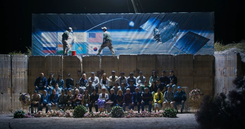 A group of people sitting on bleachers viewing a rocket launch at night, while men stand on a wall above the group painting over the flags of countries.